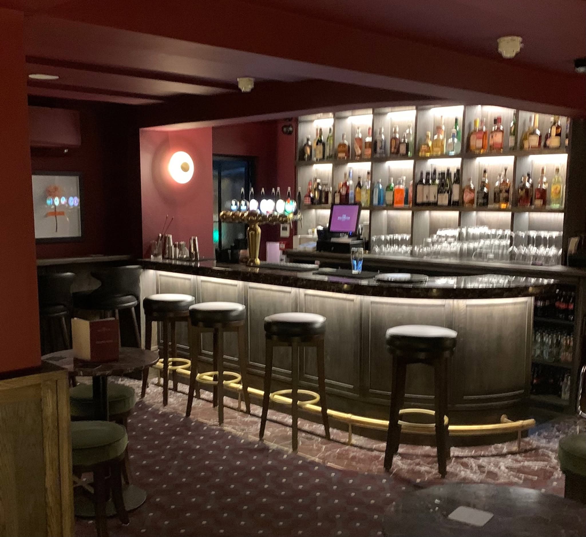 The lounge bar: oxblood walls, a backlit back bar, and a panelled counter with concealed lighting and a brass foot rail.
