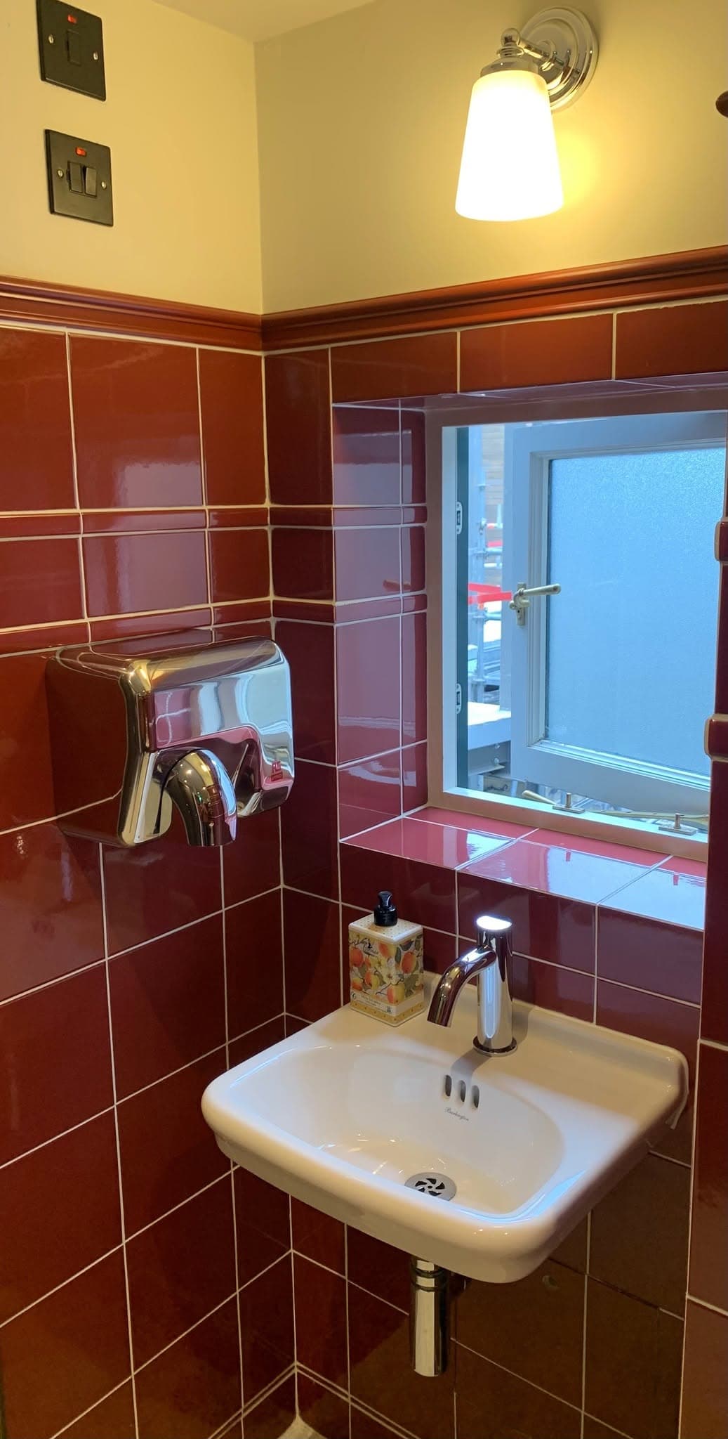 A hand-wash area in glazed oxblood tiles with a ceramic basin and brass wall light.