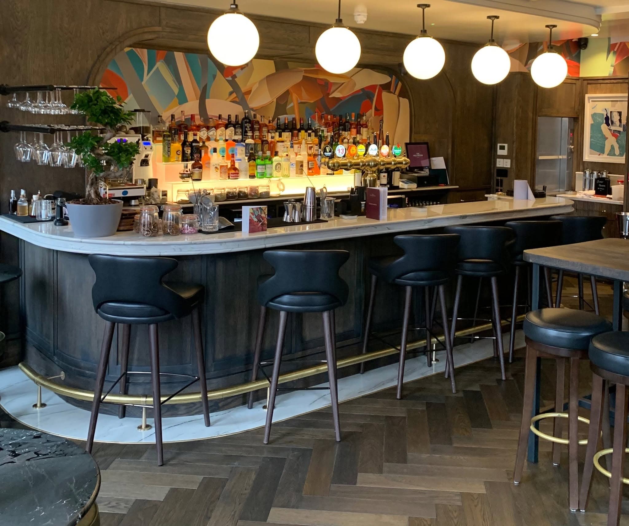 The main bar: a curved, timber-panelled counter with a marble top and brass foot rail, globe pendants overhead and a herringbone timber floor.